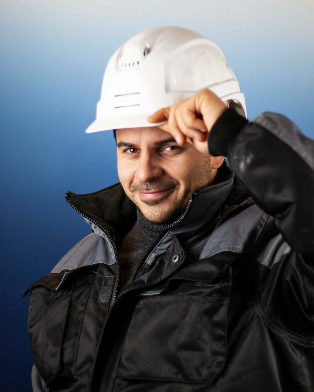 Smiling engineer in a white hard hat and dark work jacket, touching the brim of the hat